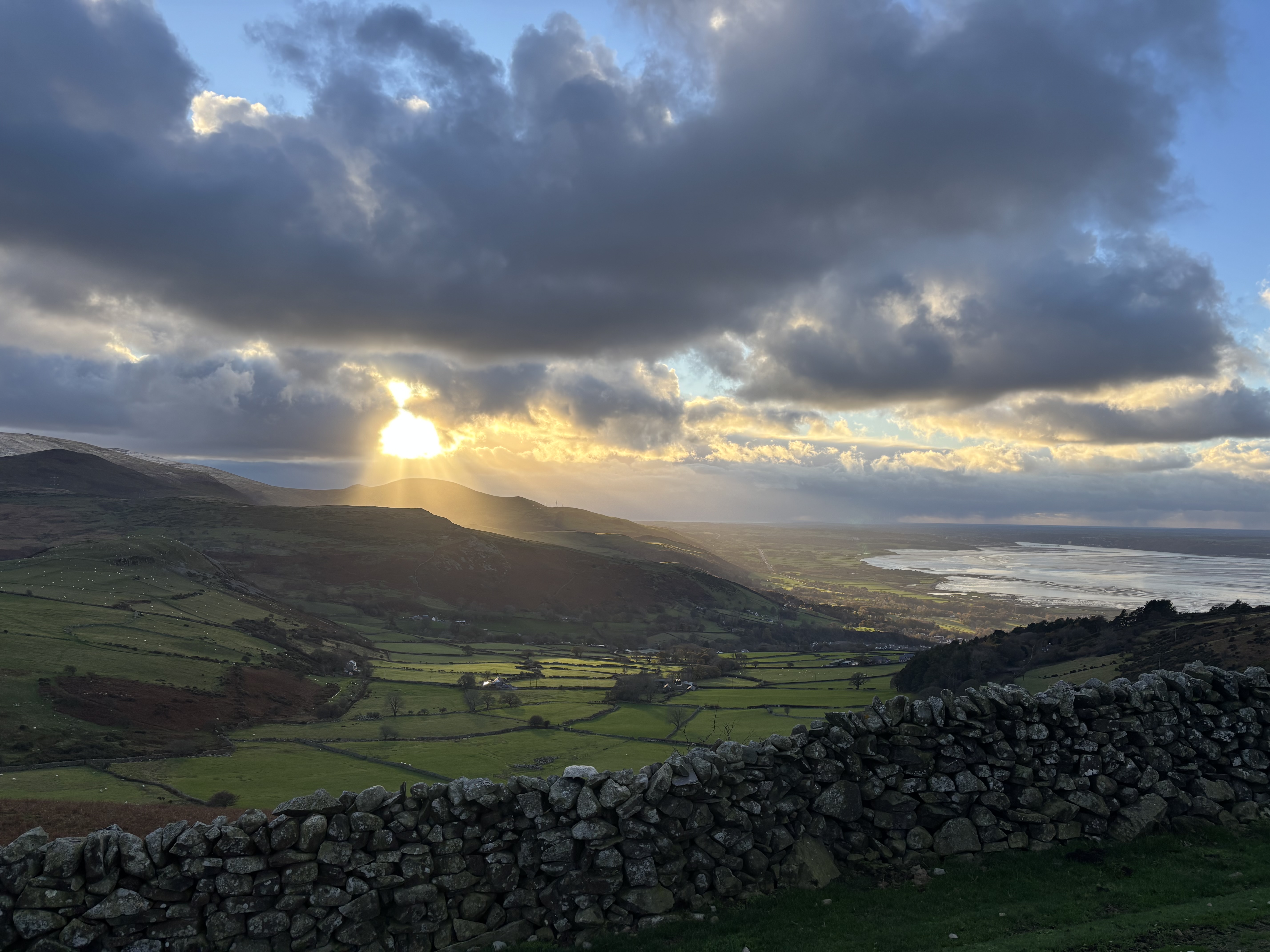 A photo across a valley near sunset. A dry stone wall is in the foreground and beyond is valley of green fields with small hills behind. The sun is shining through patchy well defined cloud, with golden rays of light.